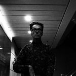 Black and white photograph of a middle-aged white man with glassed, dark hair, and a floral-patterened shirt, reflected in a large mirror